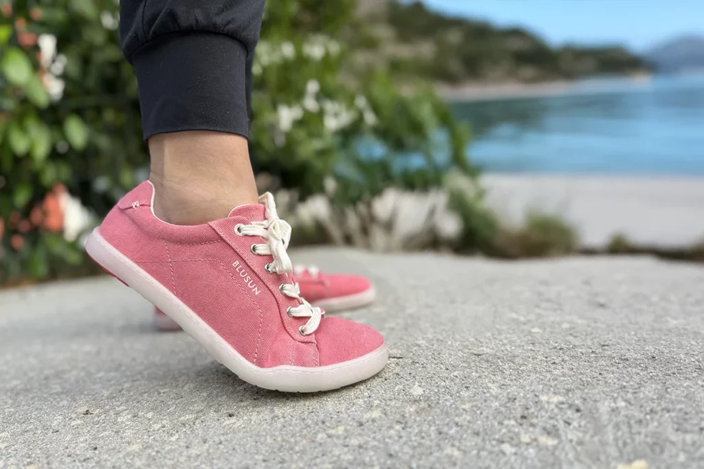 Side view of Blusun BLSN-103W pink barefoot sneakers worn on feet, with a scenic beach background