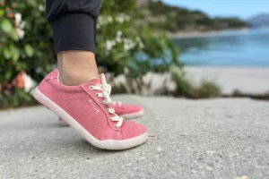Side view of Blusun BLSN-103W pink barefoot sneakers worn on feet, with a scenic beach background