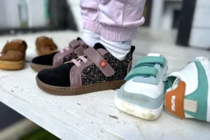 Child wearing black barefoot sneakers with pink and multicolor details, standing on a bench next to brown and teal kids’ shoes